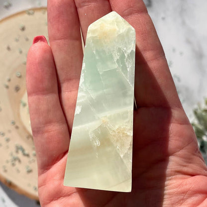 A hand holding a small, smooth, green Caribbean Calcite Obelisk with a clear, ocean-like blue underside.